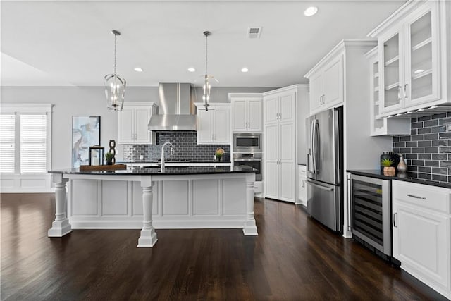 kitchen with decorative light fixtures, a center island with sink, appliances with stainless steel finishes, beverage cooler, and wall chimney range hood