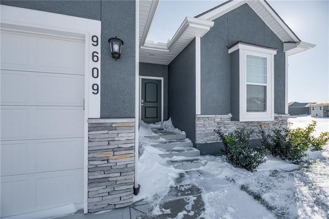 snow covered property entrance featuring stone siding, stucco siding, and a garage