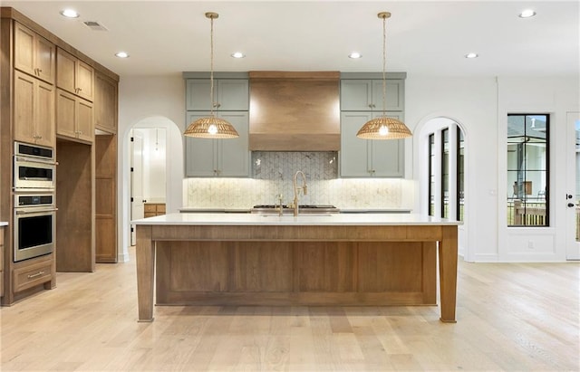 kitchen featuring an island with sink, premium range hood, backsplash, and light hardwood / wood-style floors