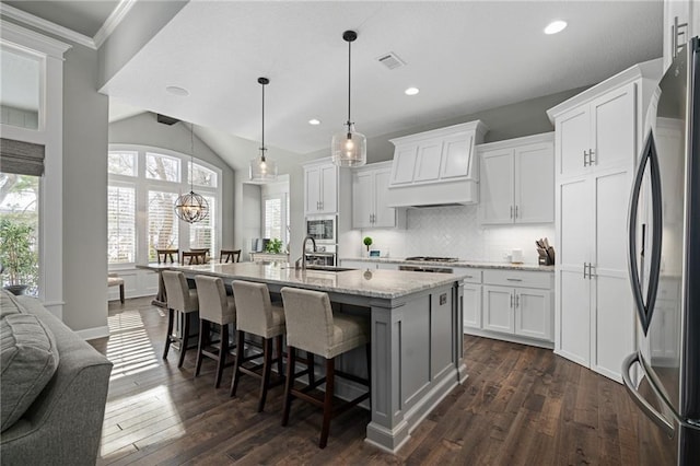 kitchen with stainless steel appliances, custom exhaust hood, white cabinets, and an island with sink