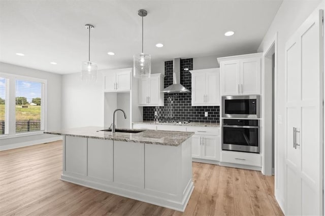 kitchen featuring sink, appliances with stainless steel finishes, wall chimney range hood, a kitchen island with sink, and white cabinets