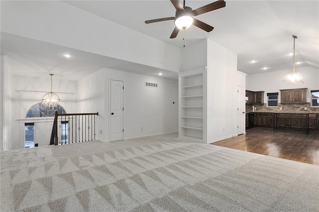 unfurnished living room with ceiling fan with notable chandelier, high vaulted ceiling, built in shelves, and dark colored carpet