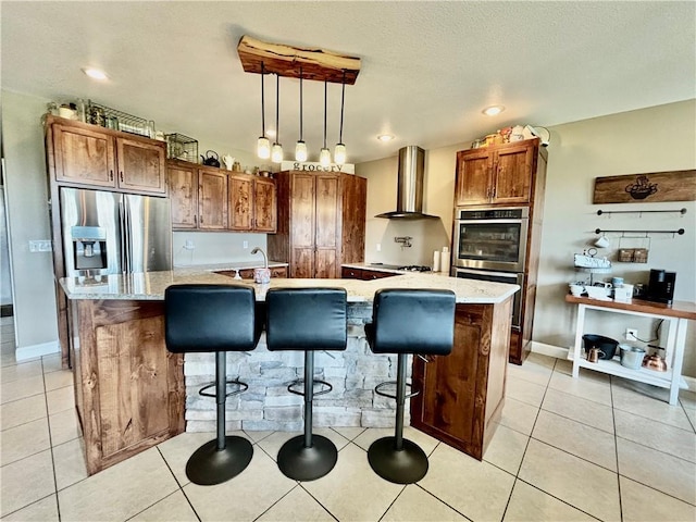 kitchen featuring stainless steel appliances, hanging light fixtures, a center island with sink, and wall chimney range hood