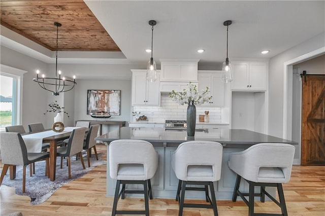 kitchen featuring a tray ceiling, a kitchen island with sink, a barn door, white cabinets, and hanging light fixtures