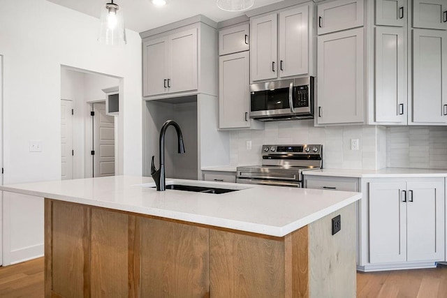 kitchen with stainless steel appliances, backsplash, an island with sink, gray cabinets, and light wood-type flooring