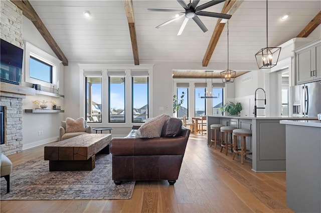 living room featuring vaulted ceiling with beams, ceiling fan with notable chandelier, a stone fireplace, and light hardwood / wood-style floors