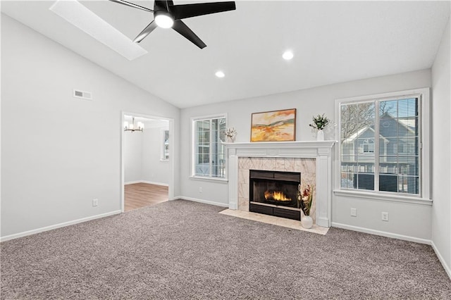 unfurnished living room featuring a premium fireplace, vaulted ceiling with skylight, ceiling fan with notable chandelier, and carpet floors