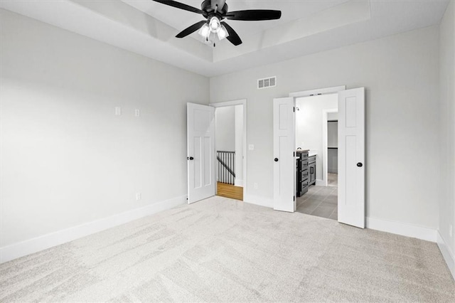 unfurnished bedroom with light carpet, a tray ceiling, ceiling fan, and ensuite bathroom