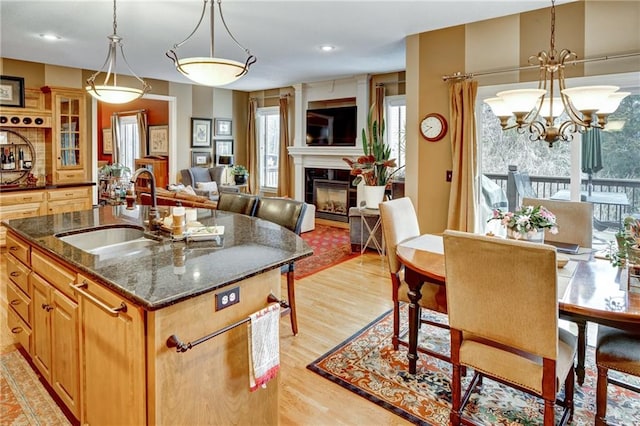 kitchen with sink, a center island with sink, decorative light fixtures, dark stone counters, and light wood-type flooring