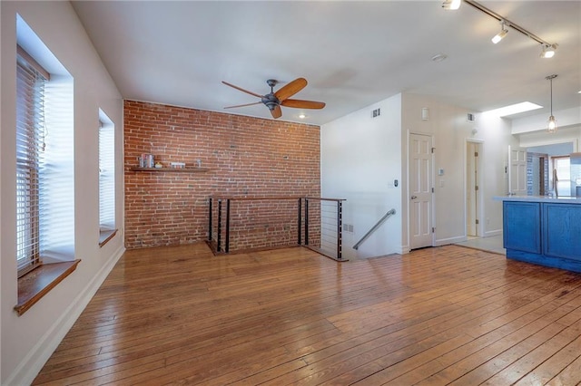 unfurnished living room with wood-type flooring, rail lighting, ceiling fan, and brick wall