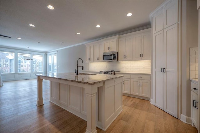 kitchen featuring a center island with sink, white cabinets, light wood-style flooring, stainless steel microwave, and a sink