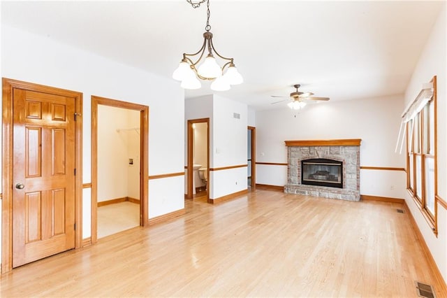 unfurnished living room featuring a stone fireplace, light wood-style flooring, ceiling fan with notable chandelier, visible vents, and baseboards