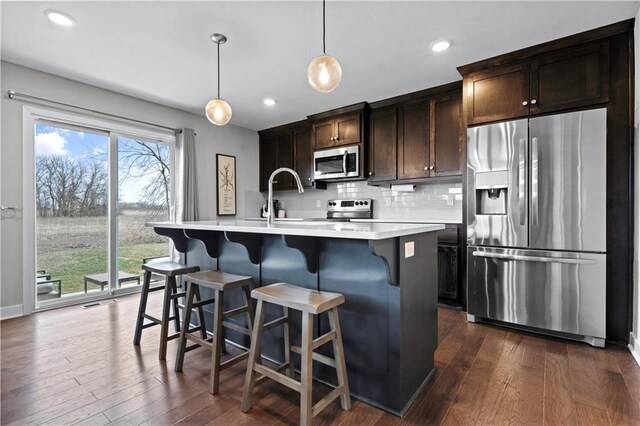 kitchen with a kitchen island with sink, stainless steel appliances, hanging light fixtures, light countertops, and dark brown cabinets