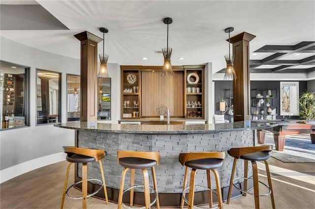 bar featuring dark stone counters, beamed ceiling, sink, hanging light fixtures, and decorative columns