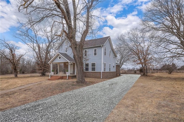 view of front of house featuring brick siding, covered porch, gravel driveway, and roof with shingles