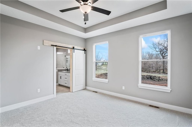 unfurnished bedroom with a raised ceiling, ceiling fan, a barn door, ensuite bath, and light colored carpet