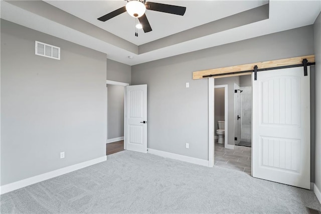 unfurnished bedroom with ensuite bathroom, a barn door, a tray ceiling, ceiling fan, and light carpet