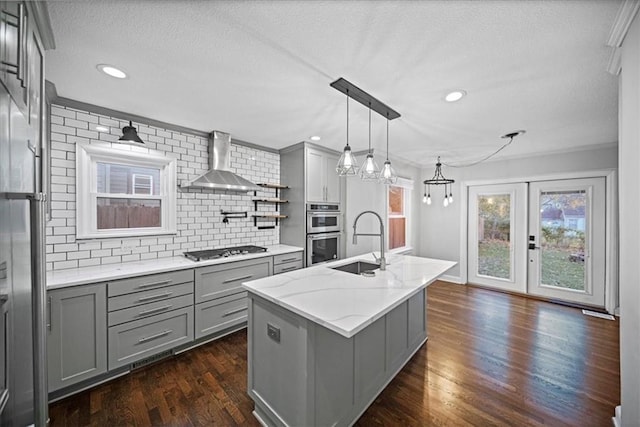 kitchen featuring wall chimney exhaust hood, an island with sink, decorative backsplash, gray cabinetry, and appliances with stainless steel finishes