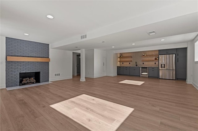 unfurnished living room with wine cooler, a brick fireplace, indoor wet bar, and light wood-type flooring