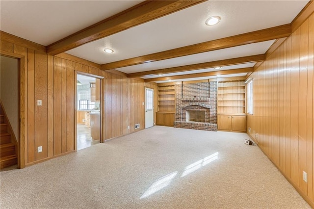 unfurnished living room featuring wooden walls, beamed ceiling, carpet flooring, a brick fireplace, and built in shelves