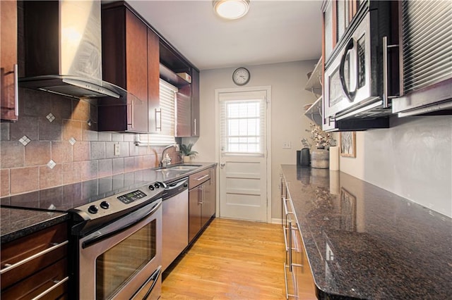 kitchen with dark stone countertops, stainless steel appliances, sink, light hardwood / wood-style flooring, and wall chimney range hood