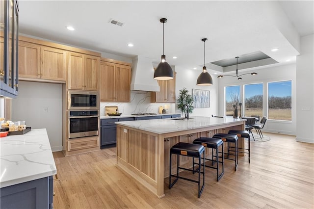 kitchen featuring light stone counters, custom exhaust hood, an island with sink, and appliances with stainless steel finishes