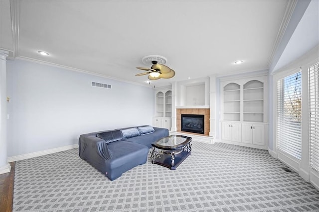 carpeted living room with ceiling fan, a tiled fireplace, built in features, and crown molding