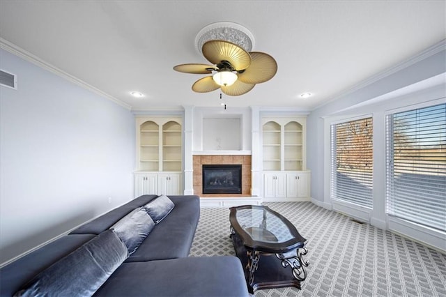 carpeted living room with ceiling fan, built in shelves, a tiled fireplace, and crown molding