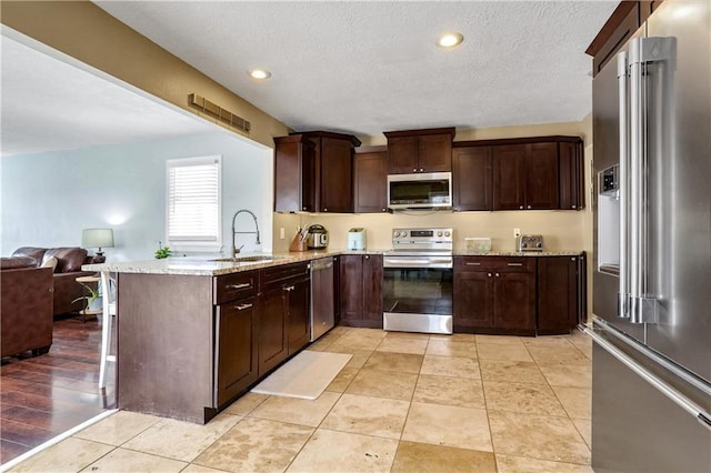 kitchen with sink, light stone counters, kitchen peninsula, stainless steel appliances, and dark brown cabinets