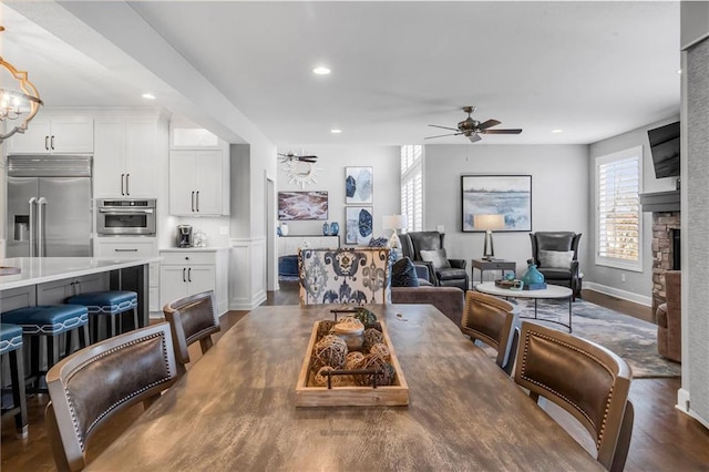 dining room with recessed lighting, ceiling fan with notable chandelier, dark wood-type flooring, a fireplace, and baseboards