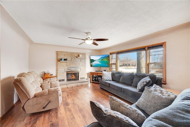 living room featuring a textured ceiling, a stone fireplace, wood finished floors, and a ceiling fan