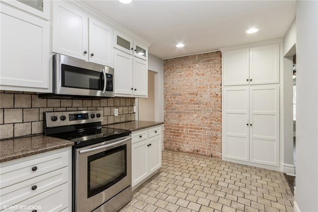 kitchen with brick wall, glass insert cabinets, dark stone countertops, stainless steel appliances, and white cabinetry