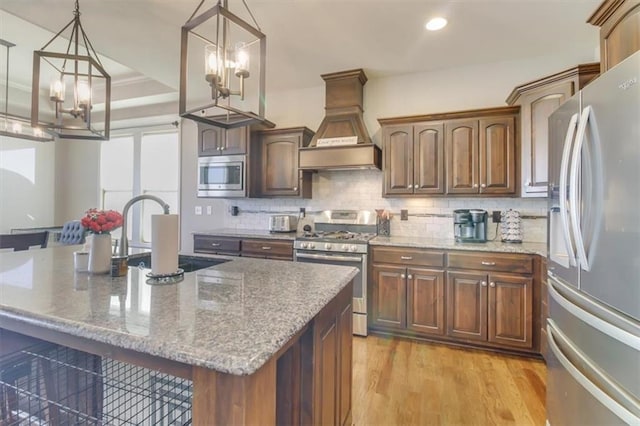 kitchen with stone countertops, decorative light fixtures, an island with sink, stainless steel appliances, and custom range hood