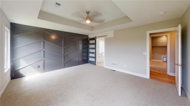 unfurnished bedroom featuring light carpet, ensuite bathroom, a barn door, ceiling fan, and a raised ceiling