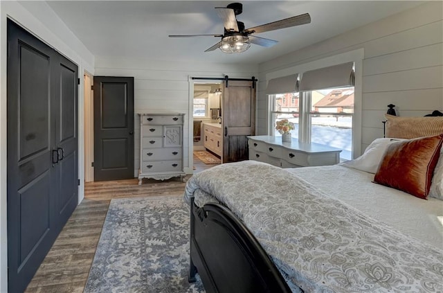 bedroom with ensuite bathroom, wooden walls, wood-type flooring, ceiling fan, and a barn door