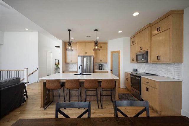 kitchen with light brown cabinets, an island with sink, pendant lighting, and appliances with stainless steel finishes