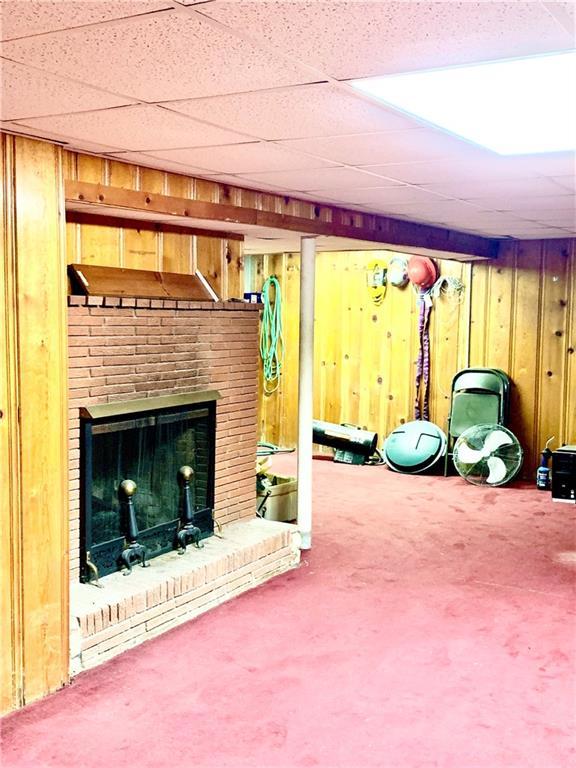 basement featuring a drop ceiling, wooden walls, a brick fireplace, and carpet floors