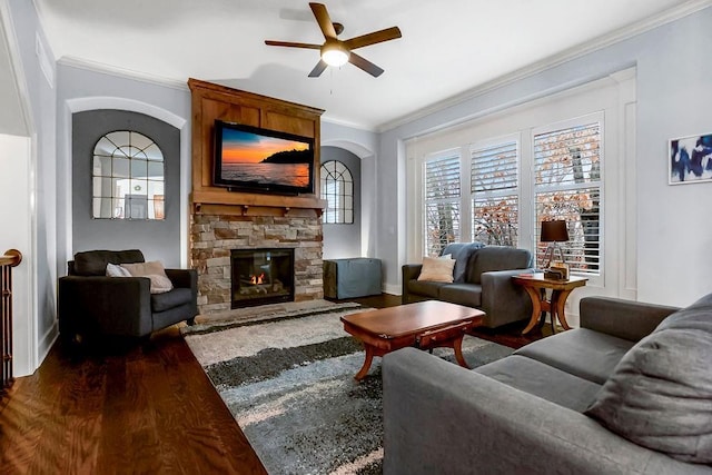 living room with dark wood-type flooring, crown molding, a stone fireplace, and a ceiling fan