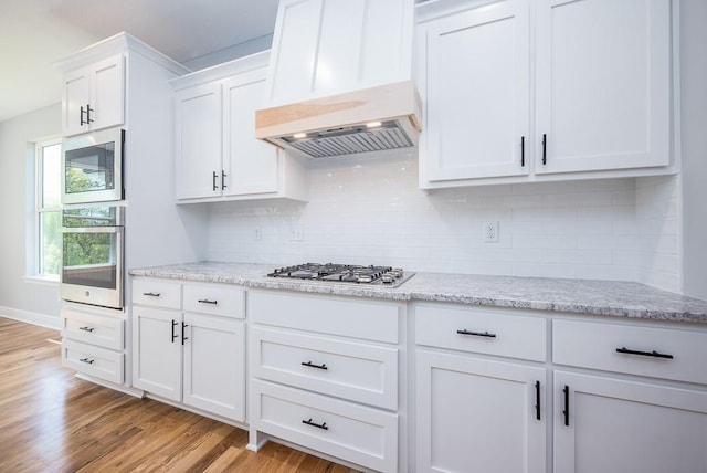 kitchen featuring light stone counters, custom range hood, appliances with stainless steel finishes, white cabinets, and light wood-type flooring