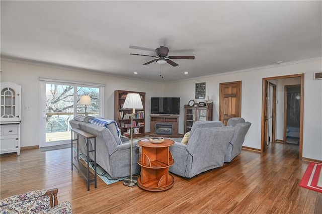 living area with baseboards, a glass covered fireplace, a ceiling fan, wood finished floors, and crown molding