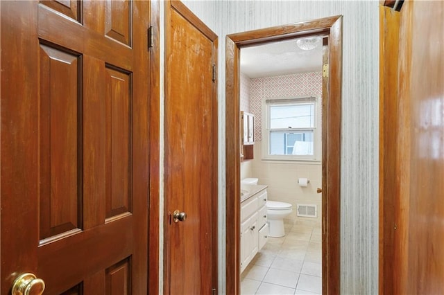 half bathroom with toilet, vanity, visible vents, tile patterned floors, and wallpapered walls