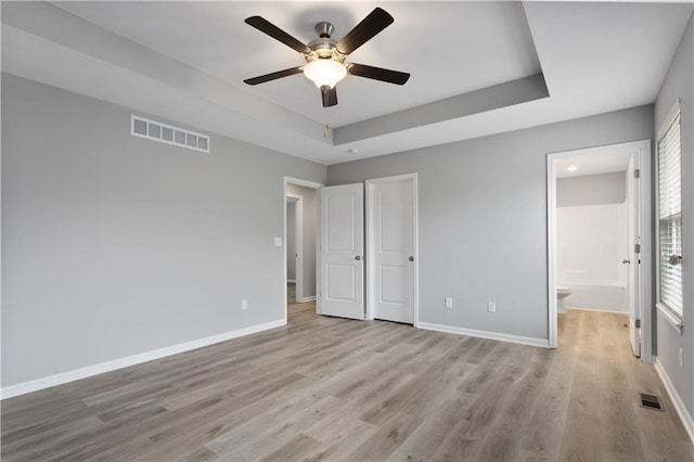 unfurnished bedroom with a raised ceiling, connected bathroom, ceiling fan, and light hardwood / wood-style floors