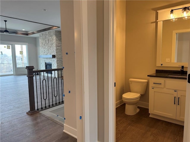 bathroom with a tray ceiling, toilet, a stone fireplace, vanity, and wood finished floors