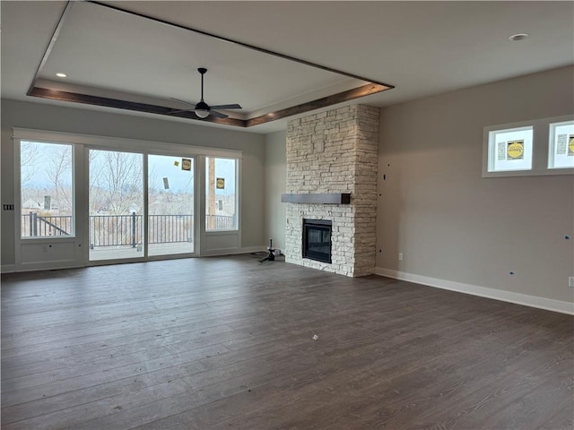 unfurnished living room with dark wood-style floors, a stone fireplace, a tray ceiling, and baseboards