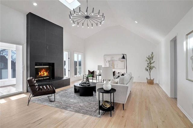 living room featuring high vaulted ceiling, a skylight, a chandelier, a tiled fireplace, and light hardwood / wood-style flooring