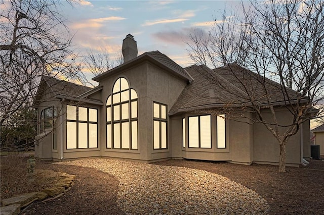 back of house at dusk featuring cooling unit, a chimney, and stucco siding