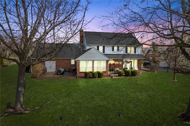 back of house at dusk with a patio area, a chimney, a lawn, and brick siding