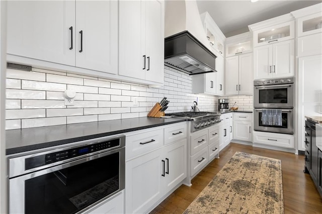 kitchen with premium range hood, white cabinetry, dark stone counters, stainless steel appliances, and backsplash