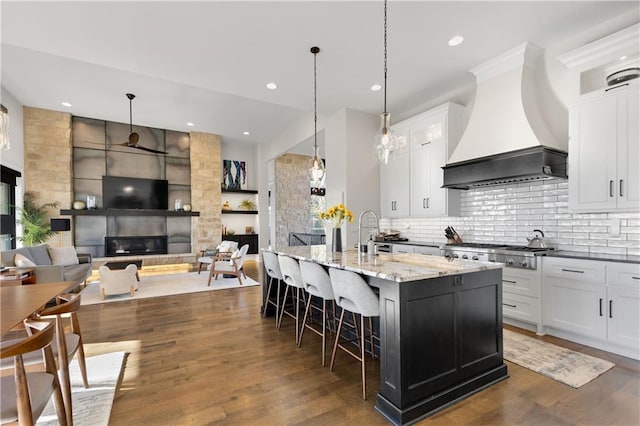 kitchen with dark stone countertops, an island with sink, custom range hood, white cabinets, and stainless steel gas cooktop