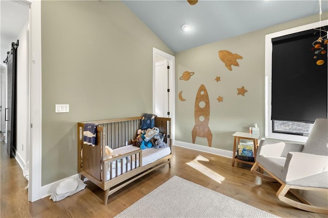 bedroom featuring a nursery area, wood-type flooring, a barn door, and vaulted ceiling